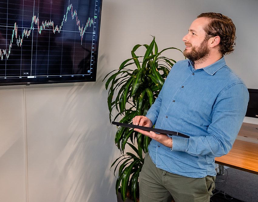 Man geeft presentatie over koersgrafieken op een scherm, met een tablet in de hand in een modern kantoor