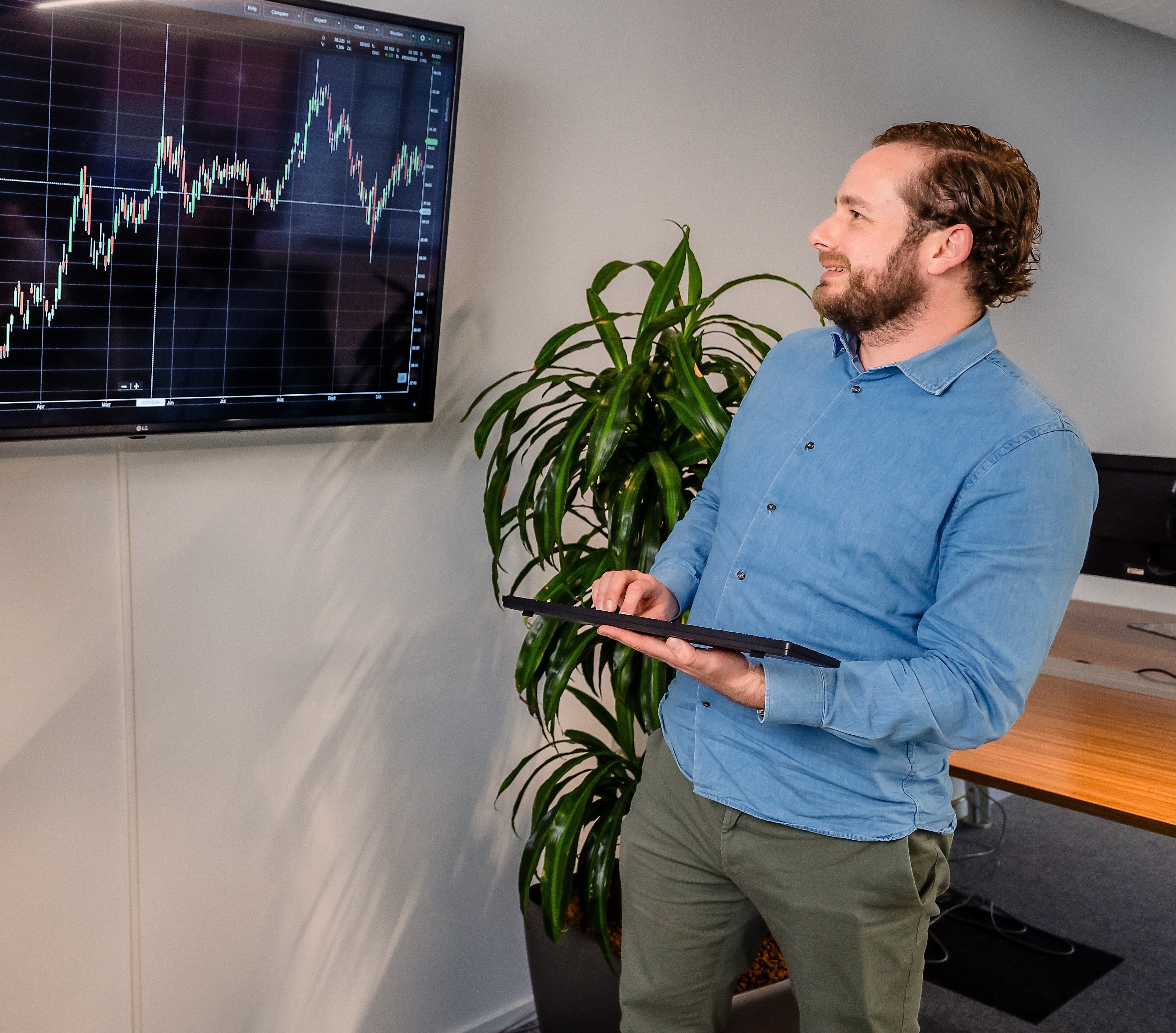 Man geeft presentatie over koersgrafieken op een scherm, met een tablet in de hand in een modern kantoor.